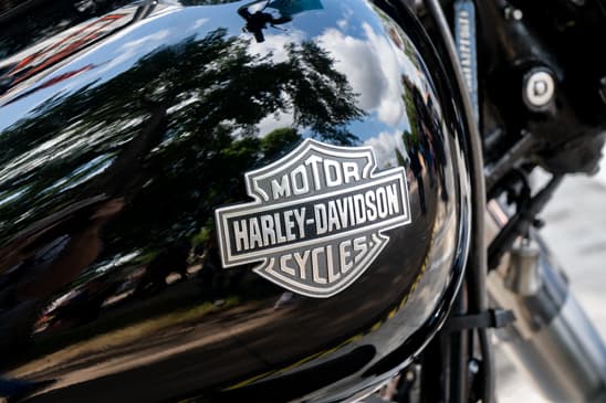 AdobeStock_282873379_Editorial_Use_Only Emblema cromato Harley-Davidson sul serbatoio nero lucido di una moto, con riflessi di alberi e cielo, ideale per test ride.