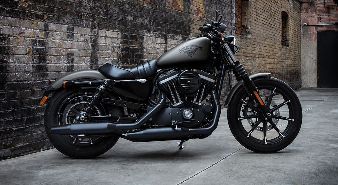 Harley-883 Harley-Davidson Iron 883 grigio opaco, motore V-twin nero e scarichi scuri, parcheggiata in un vicolo urbano.