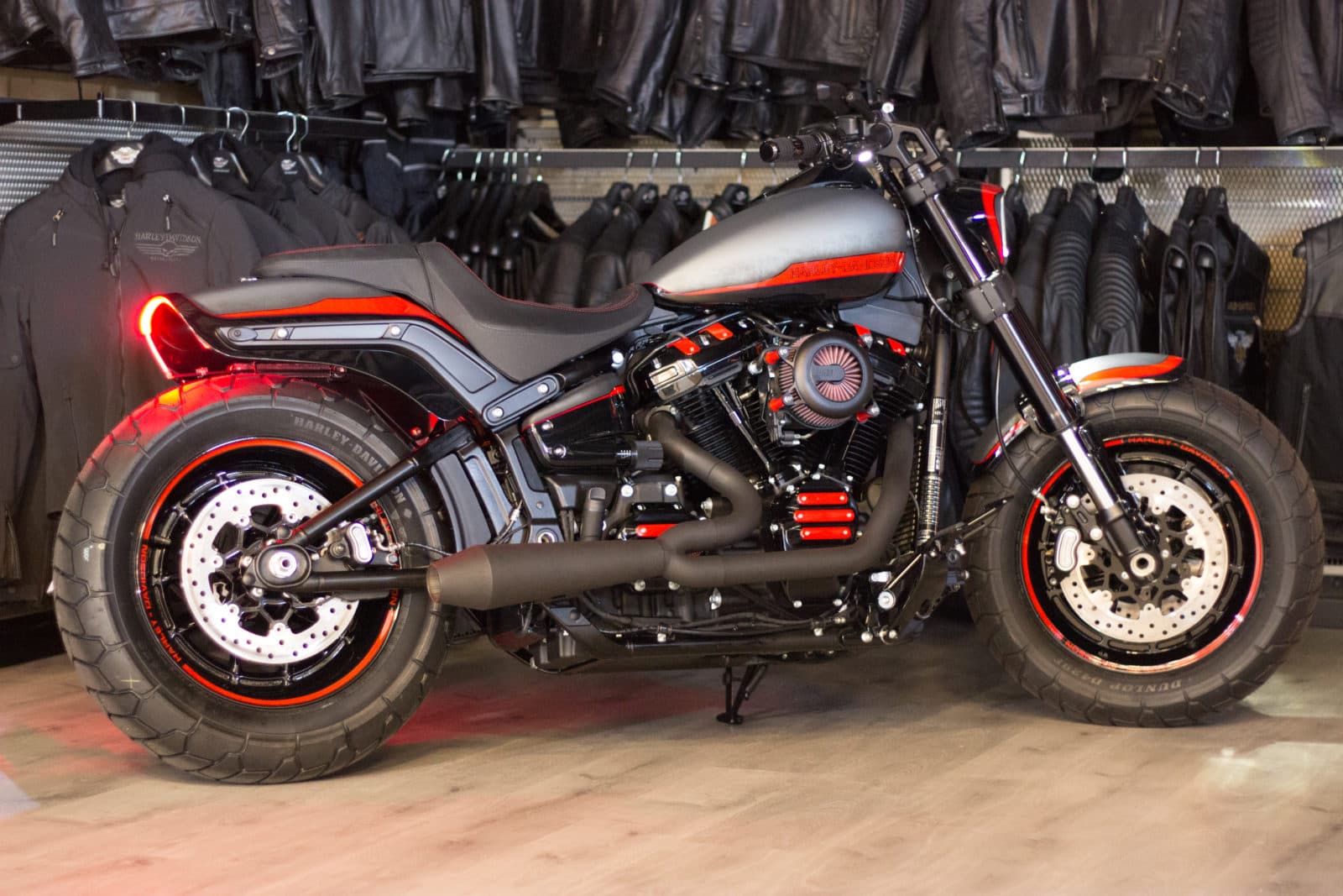 Harley-Davidon-Alba-Battle-of-the-Kings-2019-FastBob-5481.jpg Harley-Davidson customizzata, livrea nera opaca con dettagli rossi su serbatoio, motore, cerchi e fanale posteriore acceso. Esposta in concessionaria con abbigliamento Harley-Davidson.