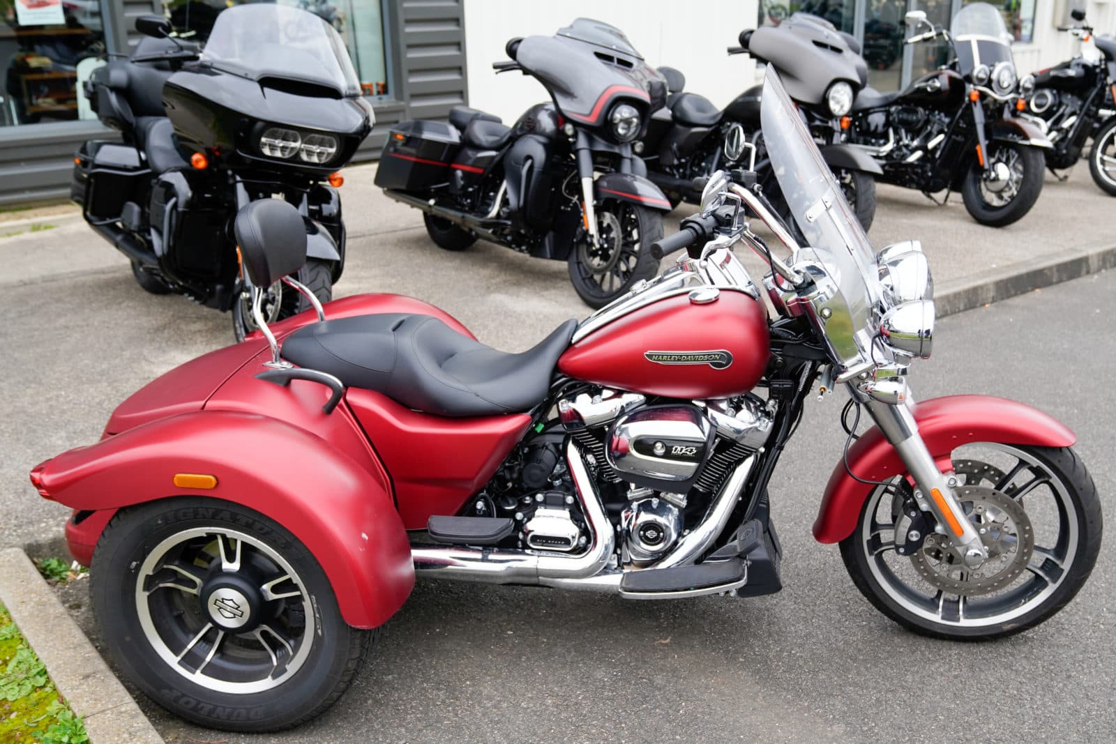 Classic Harley-Davidson trike red parked on dealership for sale retail location Harley-Davidson Trike rosso in primo piano, con modelli touring e cruiser in esposizione, suggerendo opportunità di test ride.