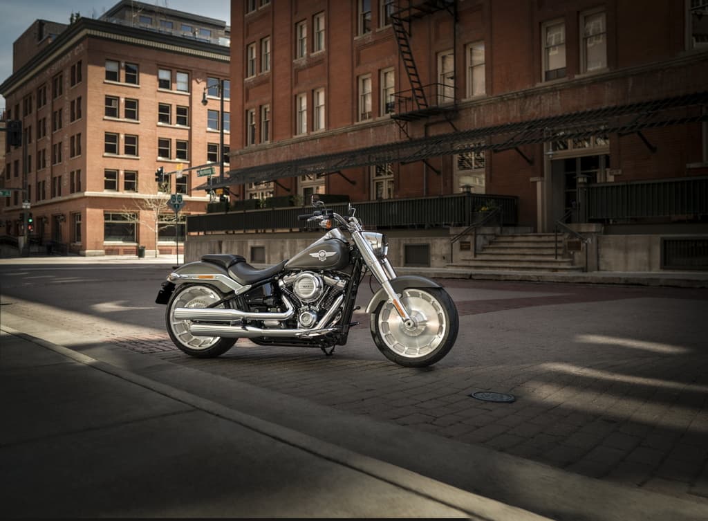 Model Year 2019 GAP Photography. INTERNATIONAL ONLY Harley-Davidson Fat Boy 114 grigio scuro parcheggiata su strada lastricata in un contesto urbano con edifici in mattoni rossi.
