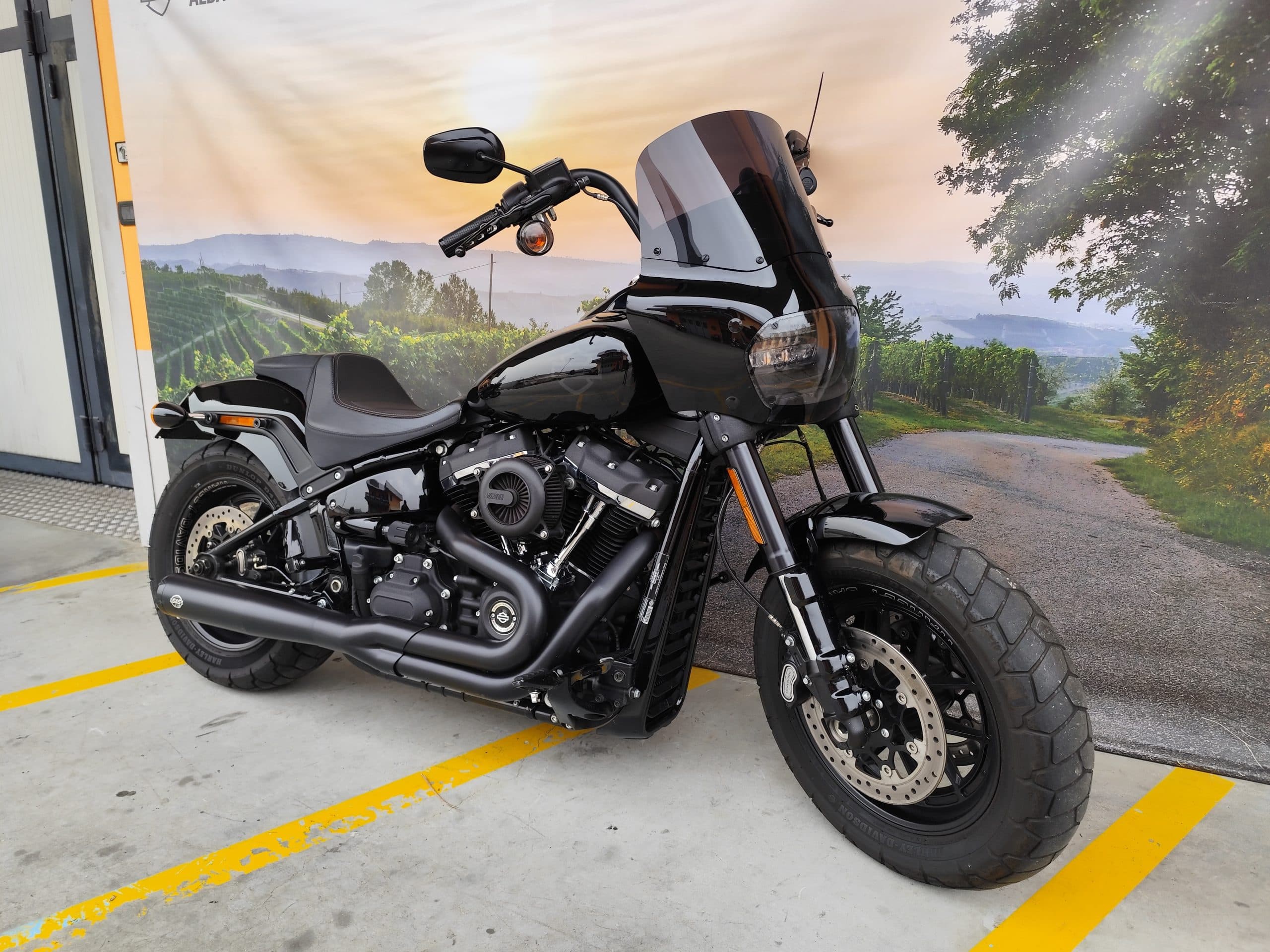 IMG_20260303_084403.jpg Harley-Davidson Fat Bob nera con cupolino, scarichi doppi e pneumatici larghi, esposta in concessionaria con sfondo paesaggistico delle Langhe.
