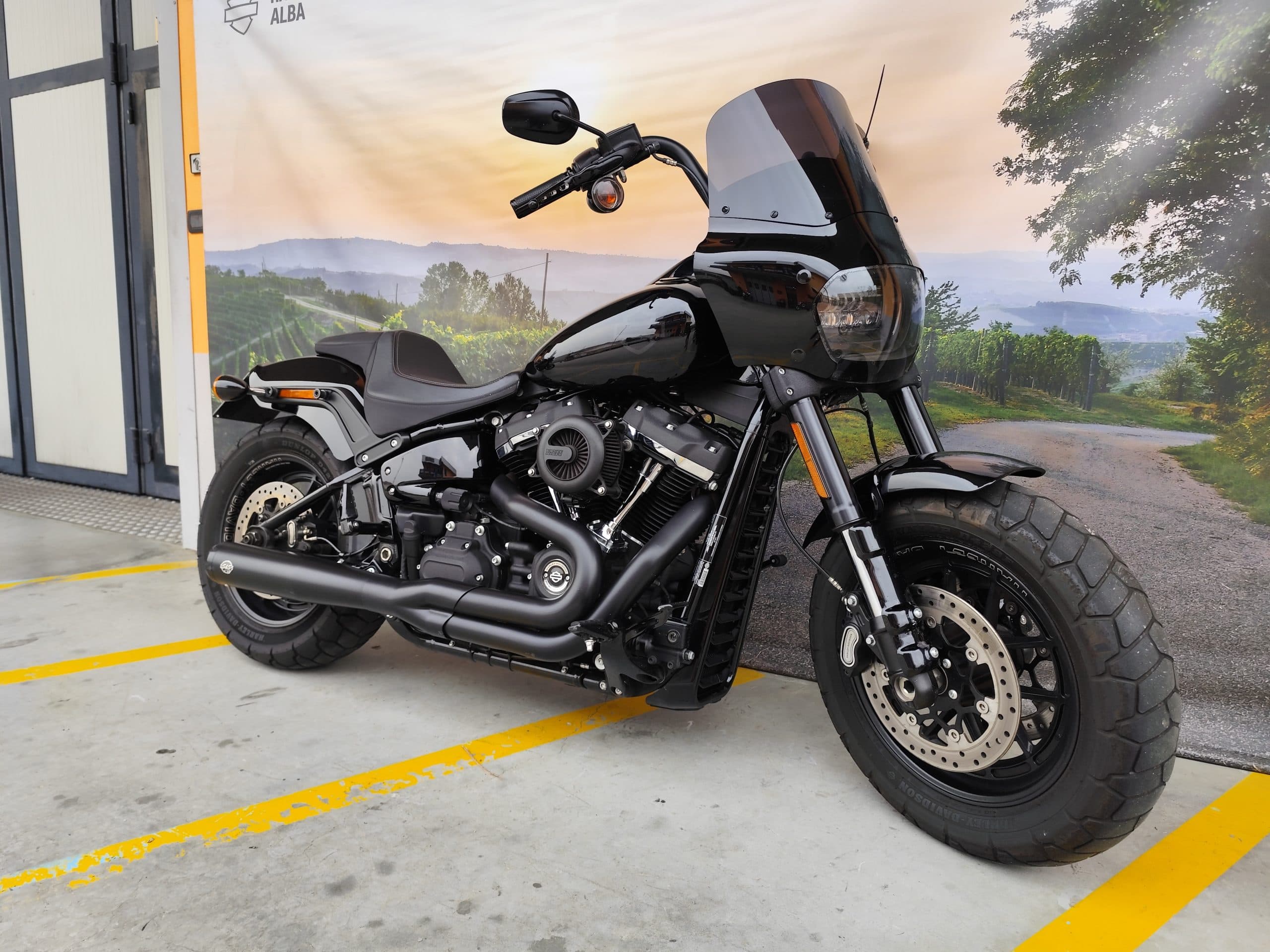IMG_20260303_084411.jpg Harley-Davidson Softail nera, con pneumatici larghi e finiture scure, parcheggiata. Sfondo banner paesaggistico di Alba. Perfetta per test ride.