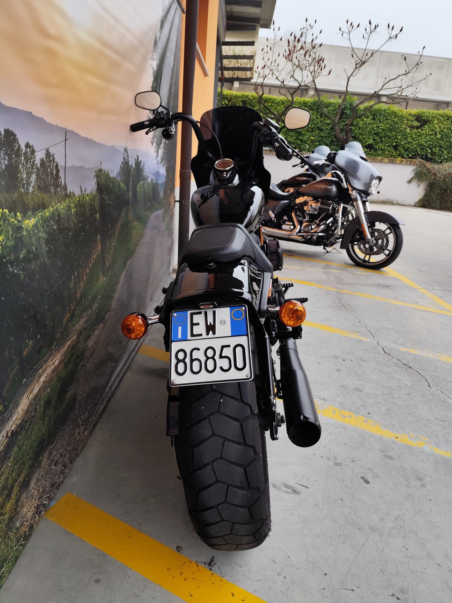 IMG_20260303_084421.jpg Harley-Davidson nera, vista posteriore, con targa italiana EW 86850, in area parcheggio. Presente altra moto Harley-Davidson grigia. Perfetta per un test ride.