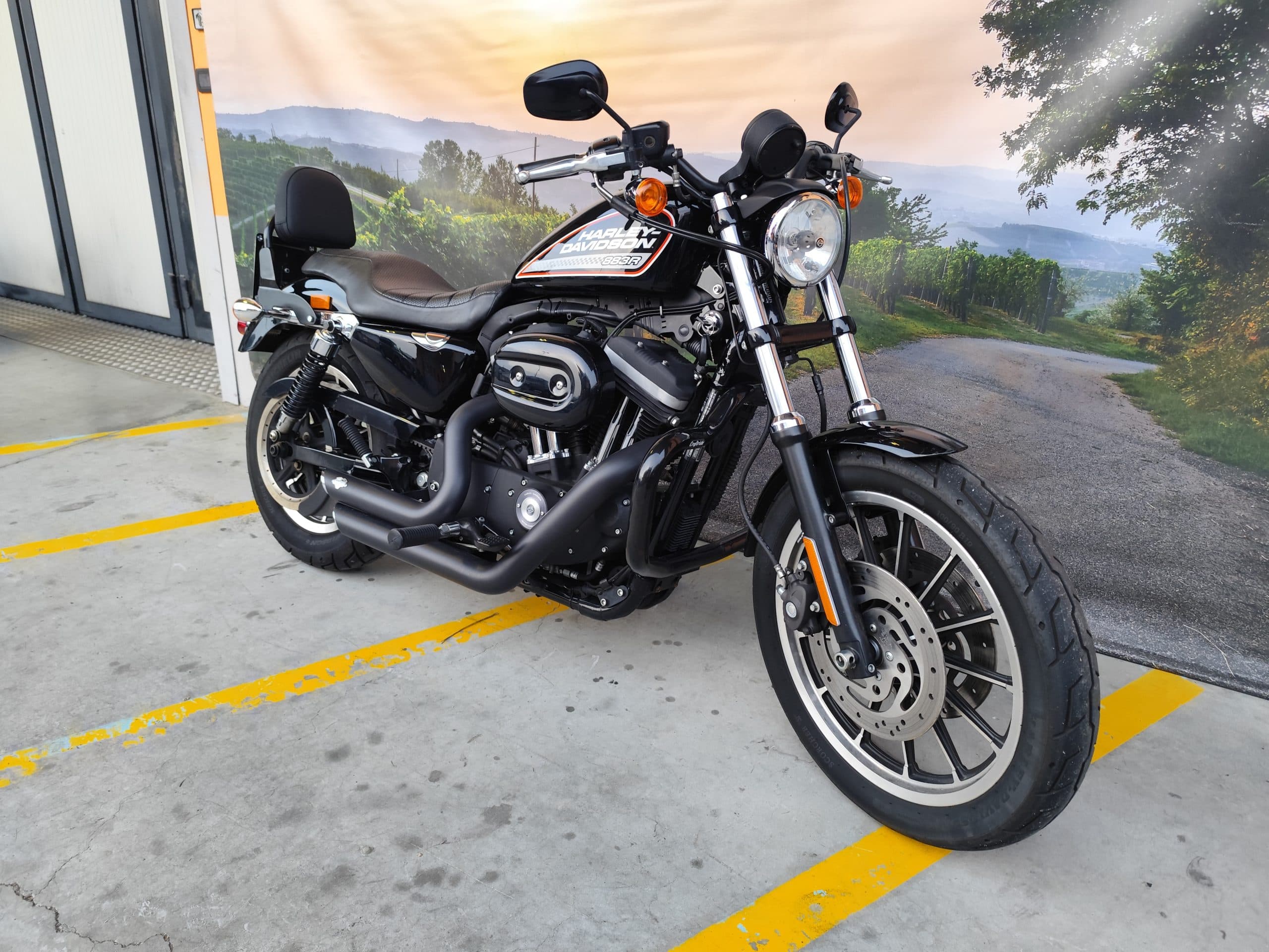 IMG_20260303_180114.jpg Harley-Davidson Sportster 883R nera, parcheggiata su cemento con strisce gialle, sfondo paesaggio collinare e vigneti. Perfetta per test ride.