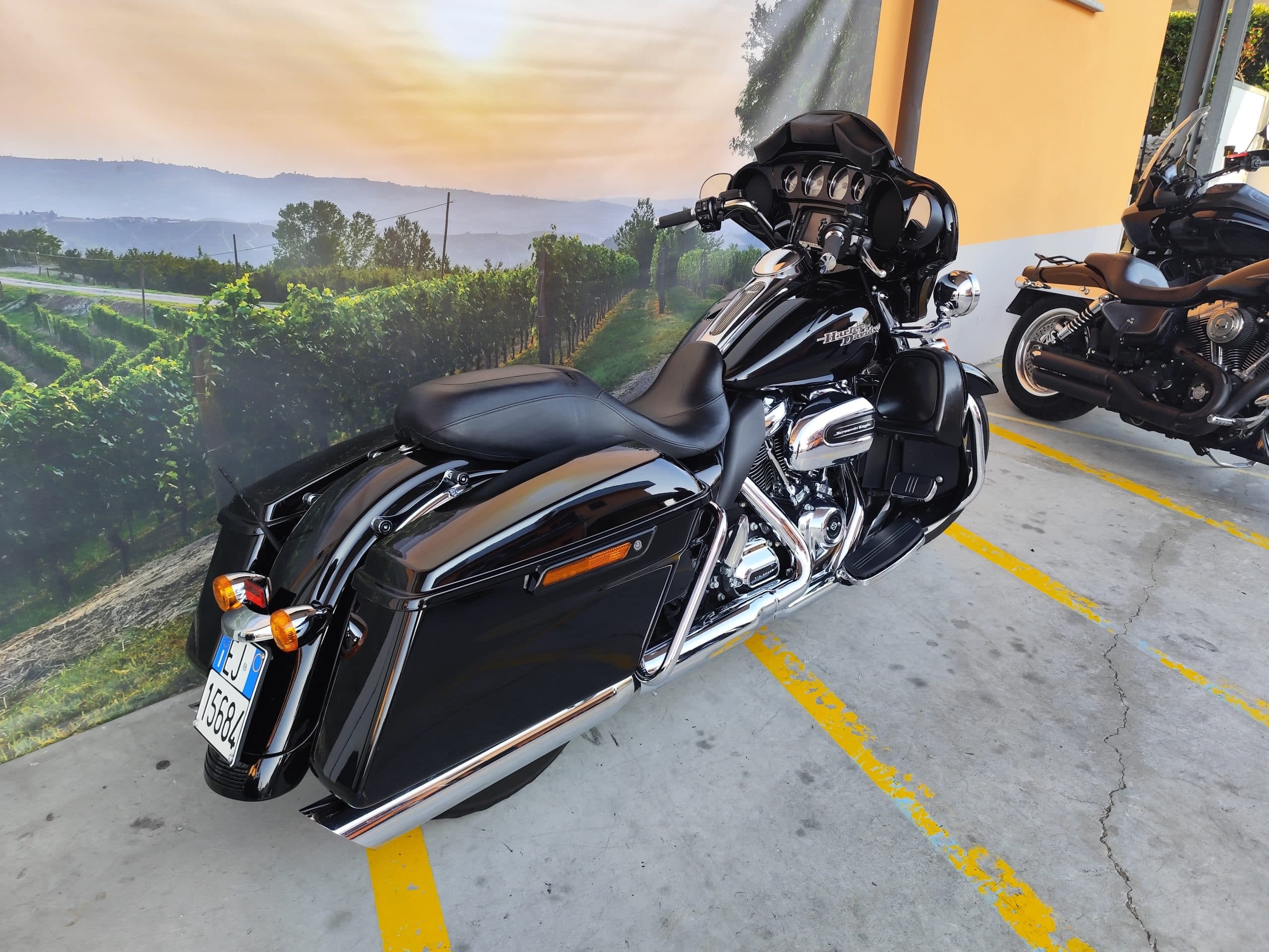 IMG_20260312_165801.jpg Harley-Davidson touring nera parcheggiata su piazzale, con sfondo murale di vigneti delle Langhe. Dettagli cromati e motore Screamin' Eagle.