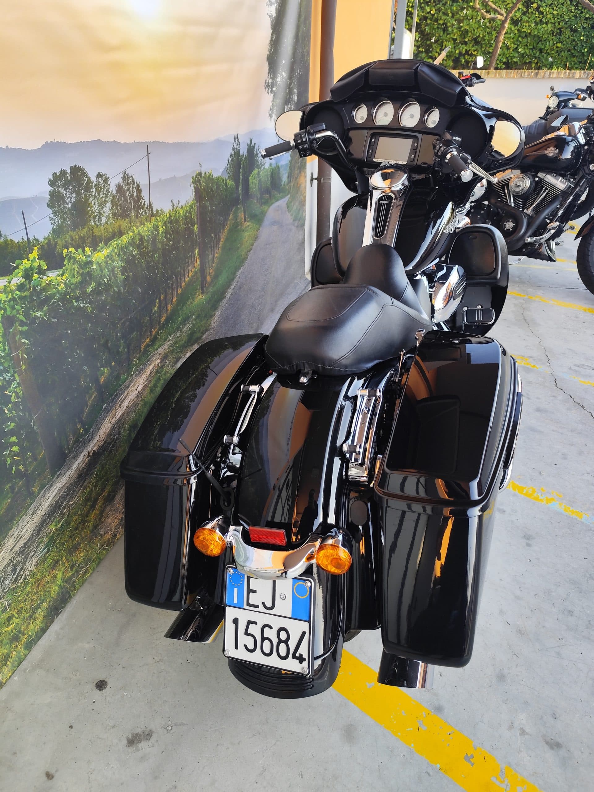 IMG_20260312_165843.jpg Harley-Davidson touring nera lucida con borse laterali e dettagli cromati, esposta davanti a un murale paesaggistico delle Langhe con vigneti e strada. Targa italiana EJ 15684.