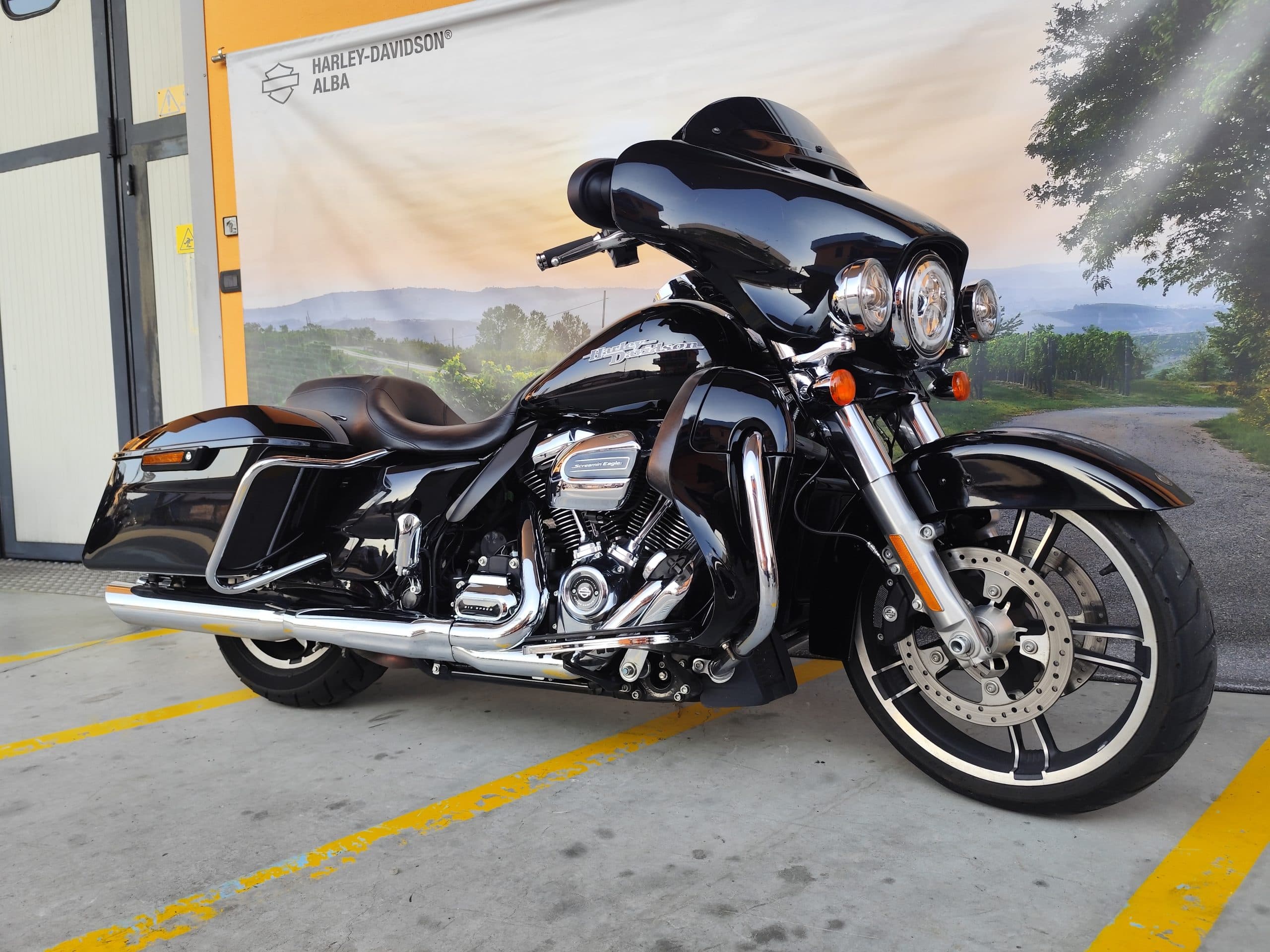 IMG_20260312_165901.jpg Harley-Davidson touring nera lucida, con dettagli cromati e motore Screamin' Eagle, parcheggiata davanti al banner di Harley-Davidson Alba. Ideale per test ride.