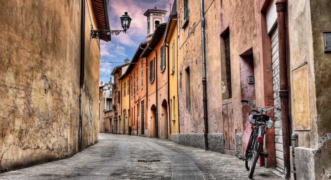 Itinerari Vicolo acciottolato con edifici storici dai colori pastello e intonaco scrostato, lampione in ferro battuto e bicicletta parcheggiata.