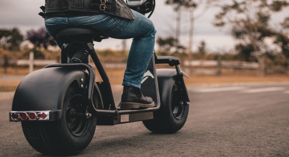 Scooter Rider con gilet in pelle, jeans e stivali alla guida di uno scooter elettrico nero. Dettaglio del veicolo con pneumatici fat e luci posteriori. Abbig