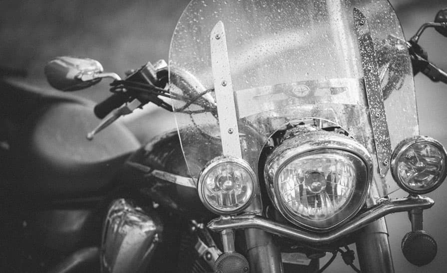 Scvino_sett_in-moto-sotto-la-pioggia Primo piano in bianco e nero del frontale di una moto Harley-Davidson, con parabrezza bagnato dalla pioggia, faro principale e luci ausiliarie cromate.