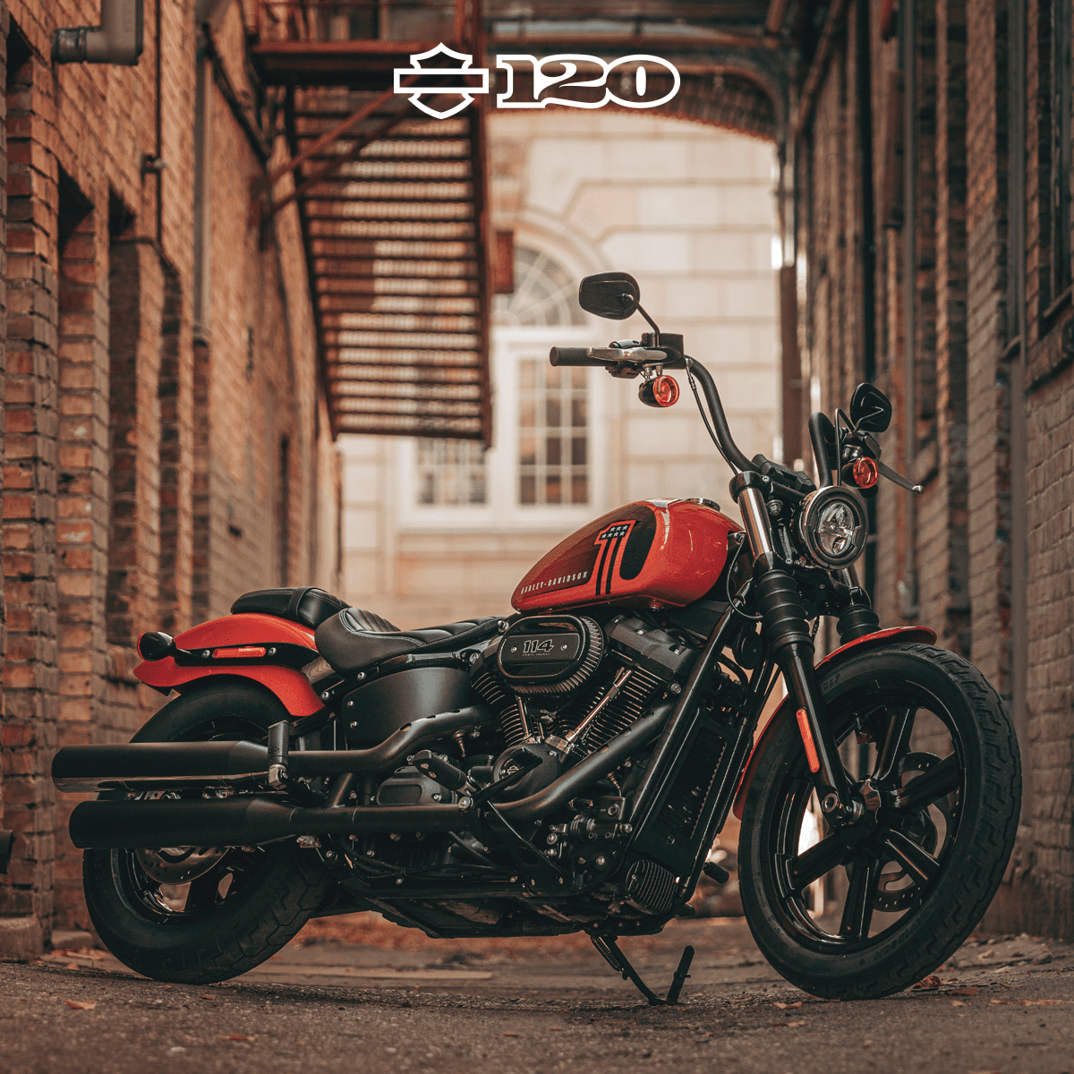 hdit-pamvoucherluglio-fbpost-4.png Harley-Davidson Softail Street Bob 114 rossa, con logo 120° anniversario, in un vicolo urbano. Moto iconica per test ride.