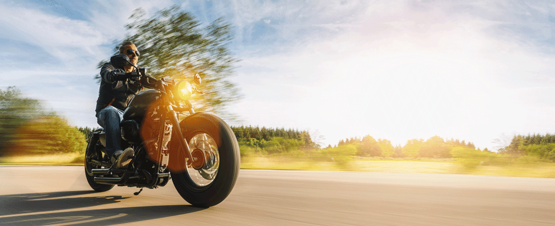 limiti-di-velocita Motociclista con giacca e occhiali da sole su moto custom in movimento, sfondo sfocato. L'emozione di un test ride con i motori