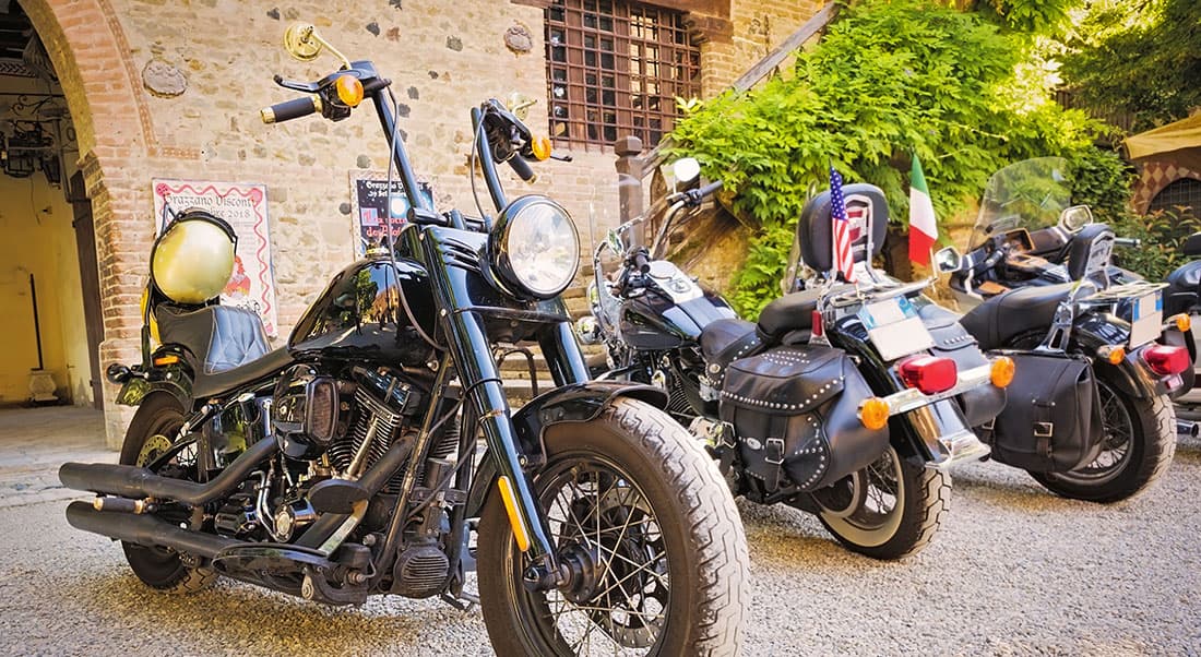 motoraduni-Harley-Davidson Moto Harley-Davidson nere parcheggiate su ghiaia, con dettagli cromati e borse laterali, in un contesto architettonico storico italiano. Visibili bandiere USA e Italia.