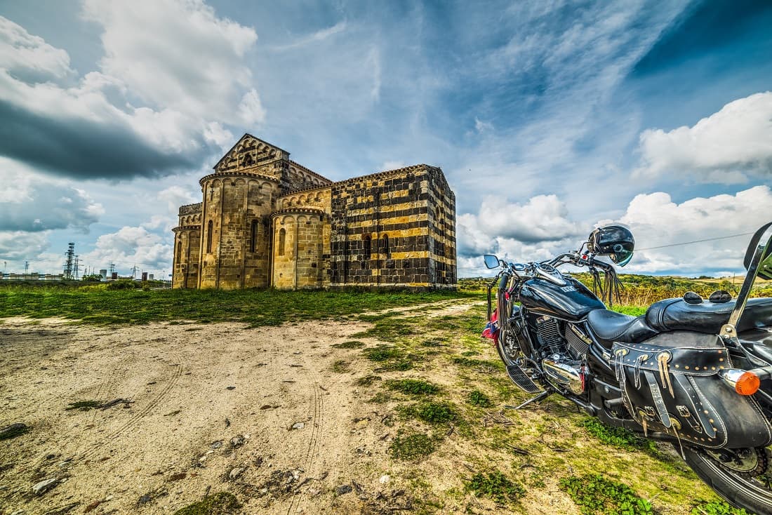 scavino-15-dicembre Harley-Davidson cruiser nera con borse in pelle, parcheggiata su sterrato davanti a una chiesa romanica. Pronta per un test ride.