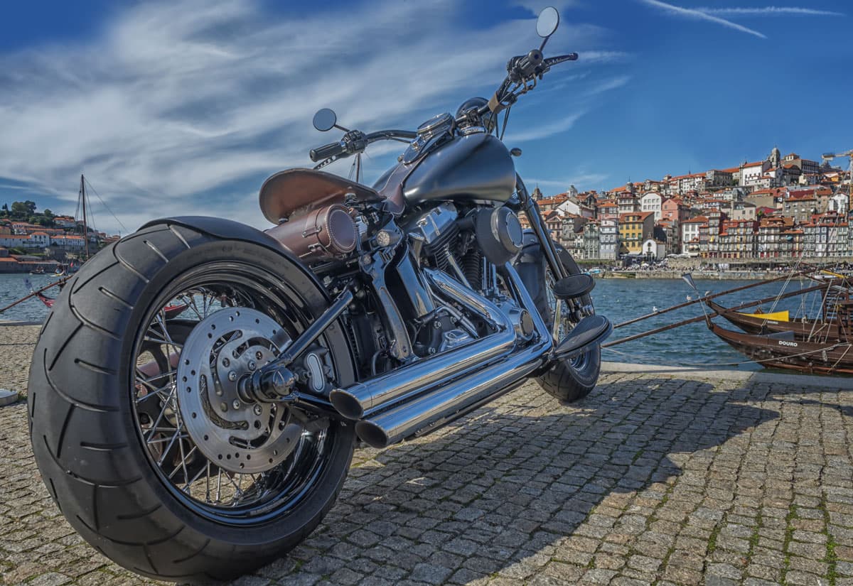scavino-2-su-3-giugno Moto custom Harley-Davidson nera opaca con scarichi cromati e sella in pelle, parcheggiata su un molo in ciottoli con vista su Porto.