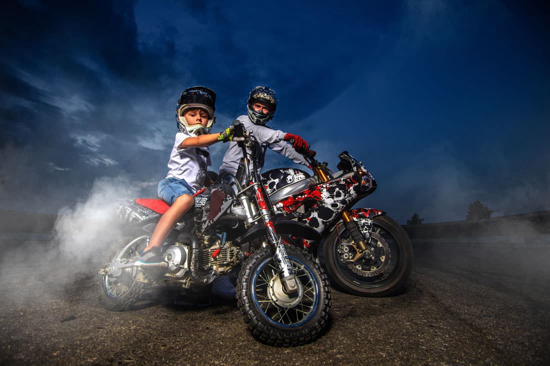 scavino-30-novembre Adulto e bambino con caschi su moto personalizzate, circondati da fumo su asfalto. Rappresenta la passione per i motori e l'abb
