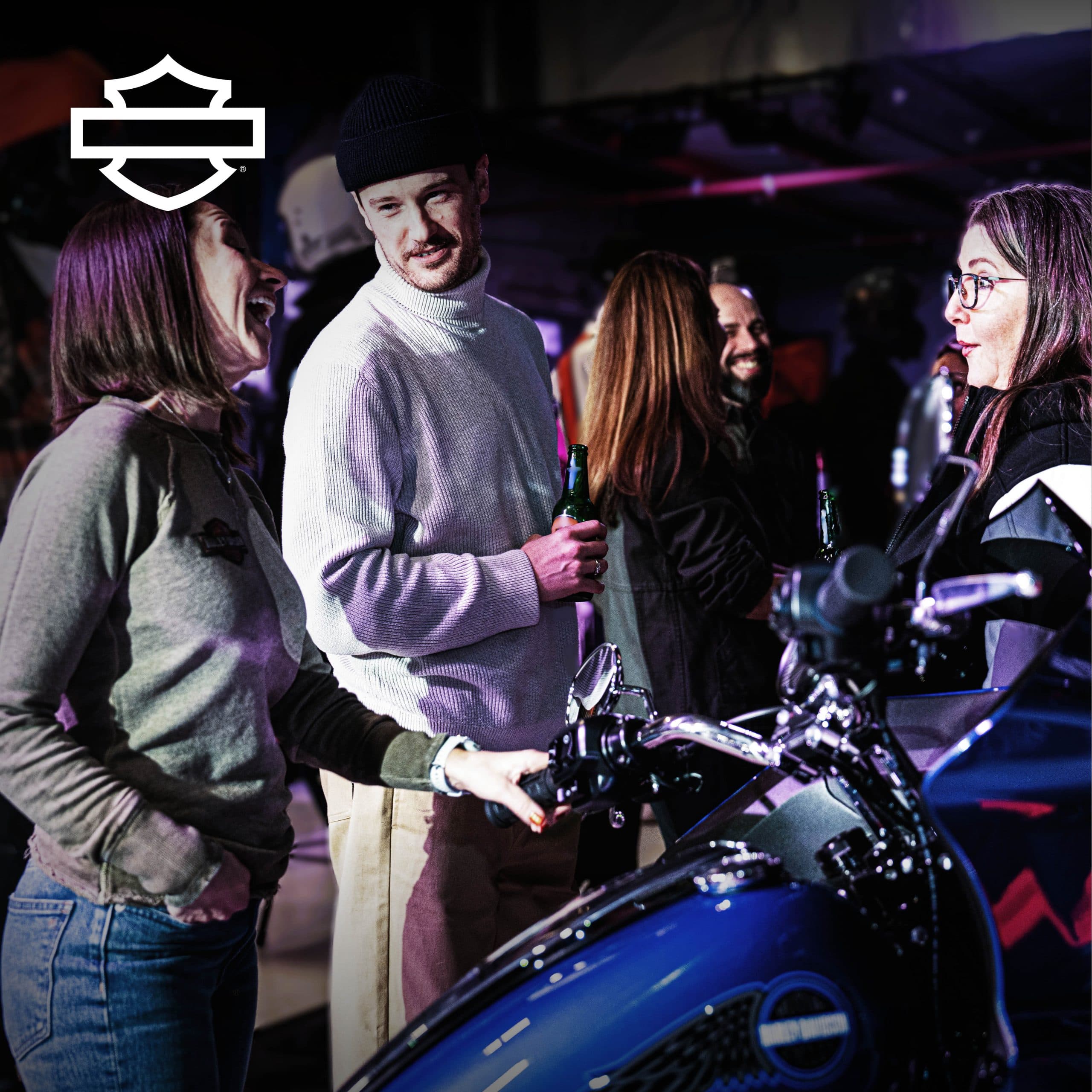 social-carosello-a-2.jpg Appassionati Harley-Davidson socializzano a un evento in concessionaria, ammirando una moto blu e l'abbigliamento del marchio.
