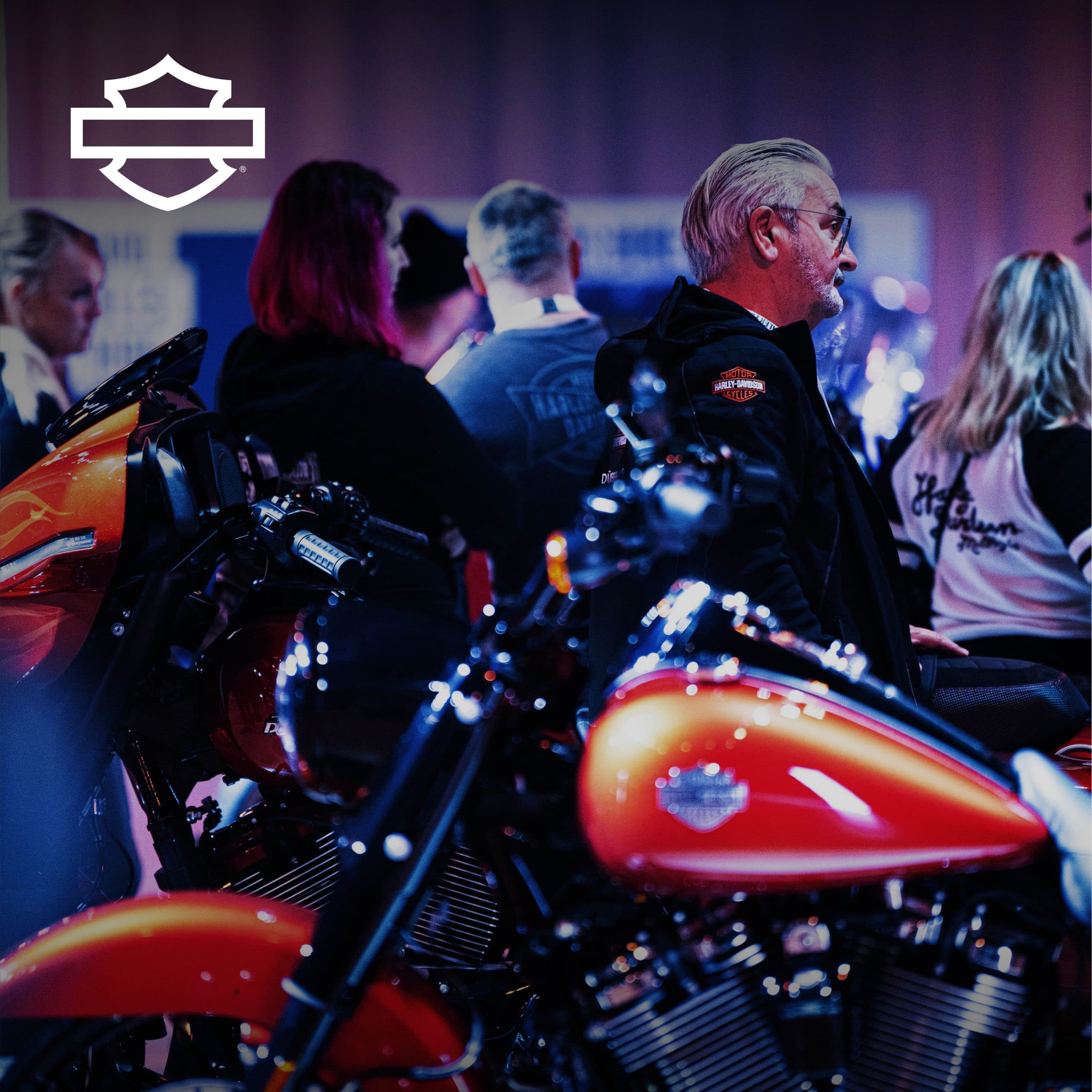 social-carosello-a-3.jpg Appassionati Harley-Davidson esaminano modelli di moto in esposizione, con focus su dettagli dei motori e abbigliamento brandizzato.