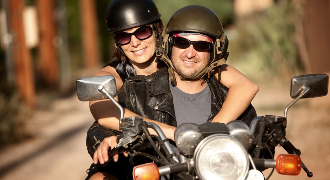 viaggiare-in-moto-in-coppia Coppia sorridente con caschi e abbigliamento in pelle su moto Harley-Davidson, pronta per un test ride o un viaggio.