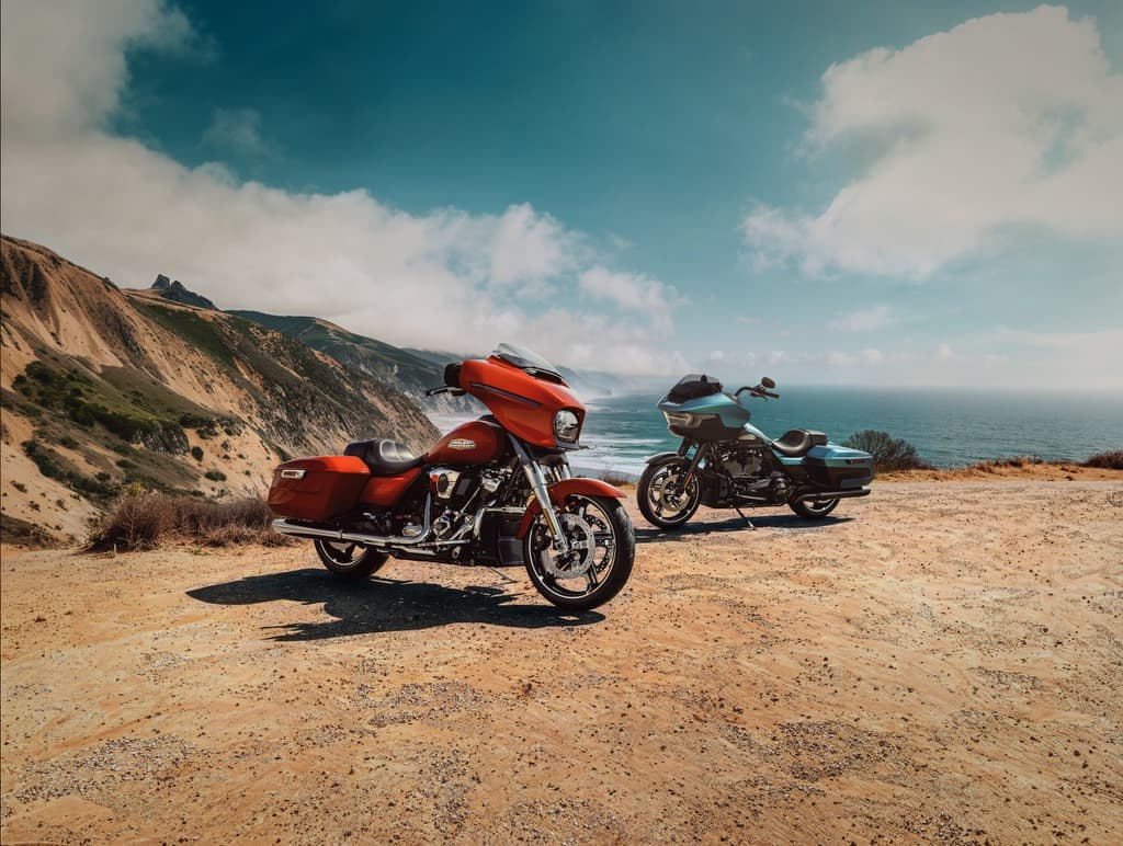 Harley-Davidson: Avventura sulla costa Due motociclette Harley-Davidson touring, una arancione e una azzurra, ferme su un sentiero sterrato con un paesaggio costiero montuoso e l'oceano sullo sfondo sotto un cielo parzialmente nuvoloso.