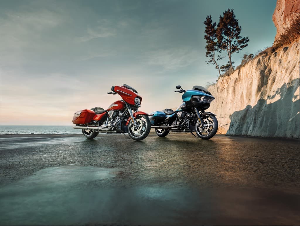 Harley-Davidson: la strada ti chiama, l’orizzonte ti aspetta Due moto Harley-Davidson touring, una arancione e una blu, parcheggiate su asfalto bagnato con riflessi, sullo sfondo mare e scogl
