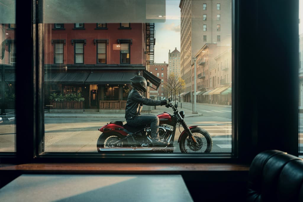 Harley-Davidson rossa in sella, pronta per la strada. Motociclista in sella a una Harley-Davidson rossa, con casco e abbigliamento tecnico, attende su strada urbana, osservato da una vetrina.