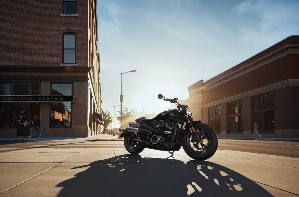 Harley-Davidson: stile urbano e libertà su due ruote Motocicletta Harley-Davidson nera, modello cruiser moderno, parcheggiata su un marciapiede urbano al tramonto. Edifici in mattoni e lunghe ombre sul selciato.