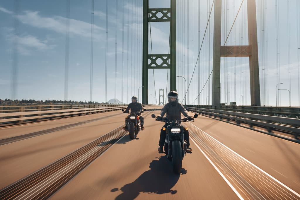 Harley-Davidson: l’emozione di un test ride. Due motociclisti con abbigliamento tecnico su moto Harley-Davidson attraversano un ponte sospeso in una giornata di sole. Esperienza di guida.