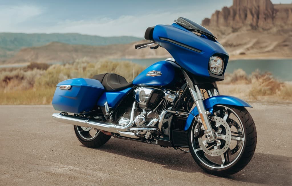 Harley-Davidson blu: l’avventura ti aspetta. Moto Harley-Davidson touring blu, con carenatura batwing e borse rigide, parcheggiata su strada. Paesaggio montano con lago sullo sfondo.