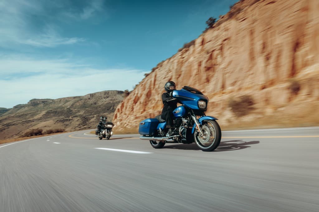 Harley-Davidson in curva: test ride su strada panoramica. Motociclista con casco e abbigliamento tecnico su Harley-Davidson blu in curva su strada asfaltata. Effetto panning per dinamismo, con altra moto in coda e paesaggio montano sfocato.