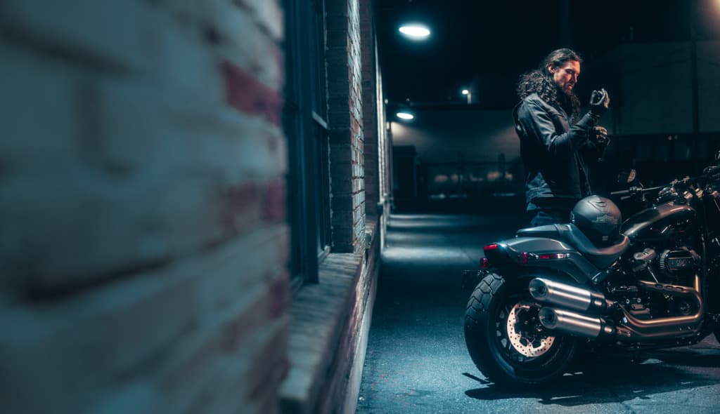 Preparazione per il test ride con Harley-Davidson Uomo con giacca e guanti in pelle che si prepara per un giro in moto Harley-Davidson nera, con casco sul sedile, in un vicolo notturno.