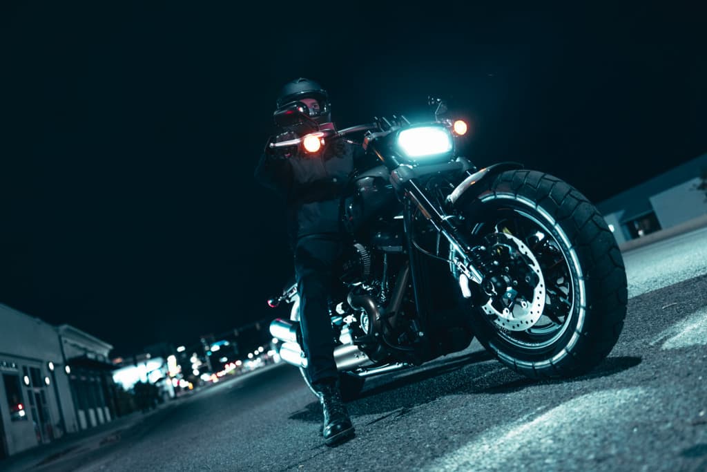 Harley-Davidson: potenza e stile nella notte. Motociclista con casco e abbigliamento tecnico su moto Harley-Davidson di notte, con faro acceso e luci di posizione arancioni, in contesto urbano.