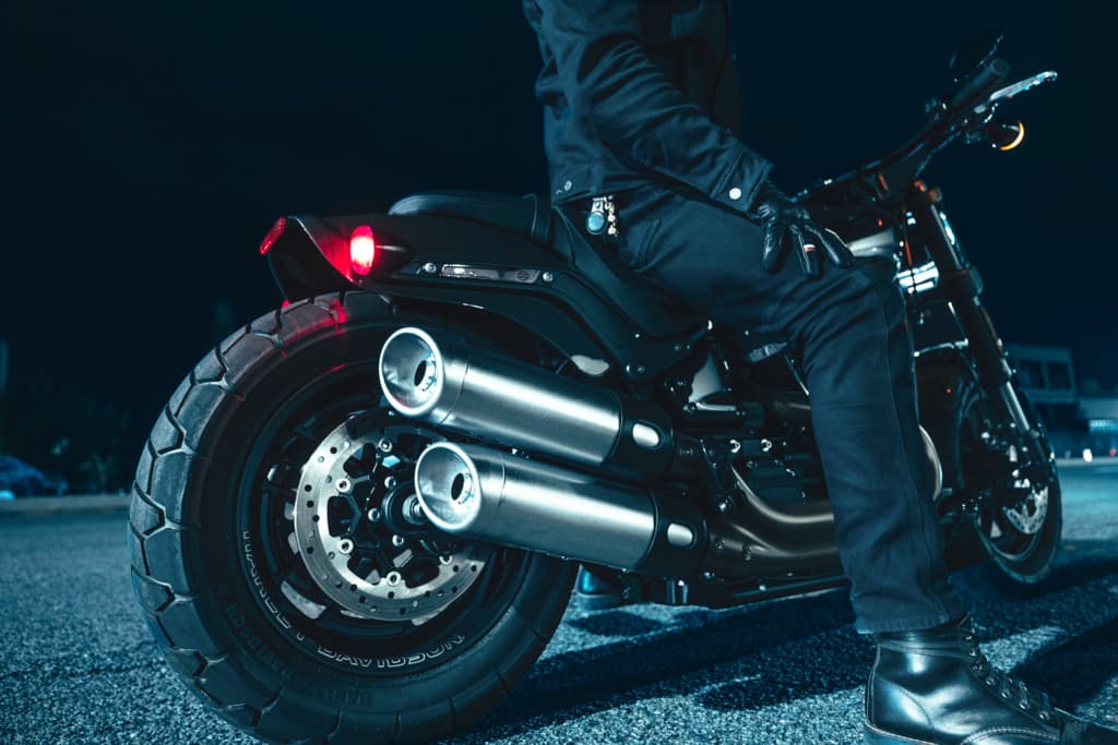 Harley-Davidson: stile e potenza Harley-Davidson con pilota in abbigliamento tecnico. Dettaglio ruota posteriore, doppio scarico e fanale rosso acceso di notte.