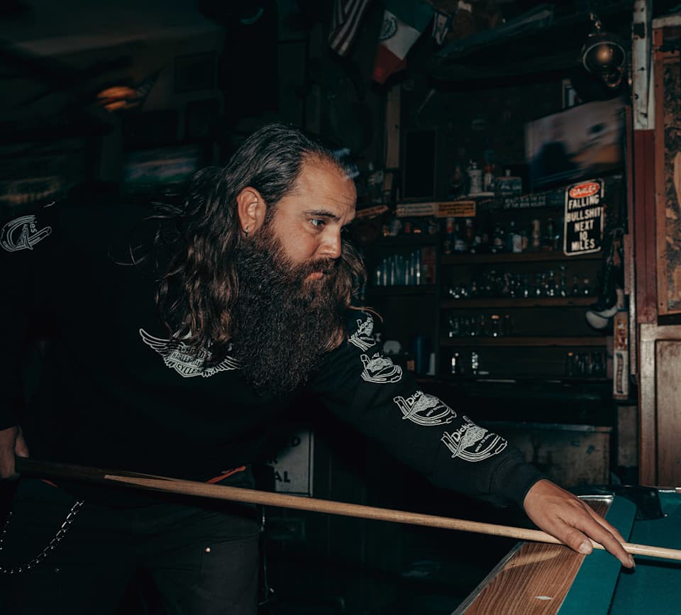 Harley-Davidson: abbigliamento e lifestyle al biliardo. Uomo con barba e capelli lunghi, indossa maglia nera Harley-Davidson con logo alato, intento a giocare a biliardo in un ambiente da pub.