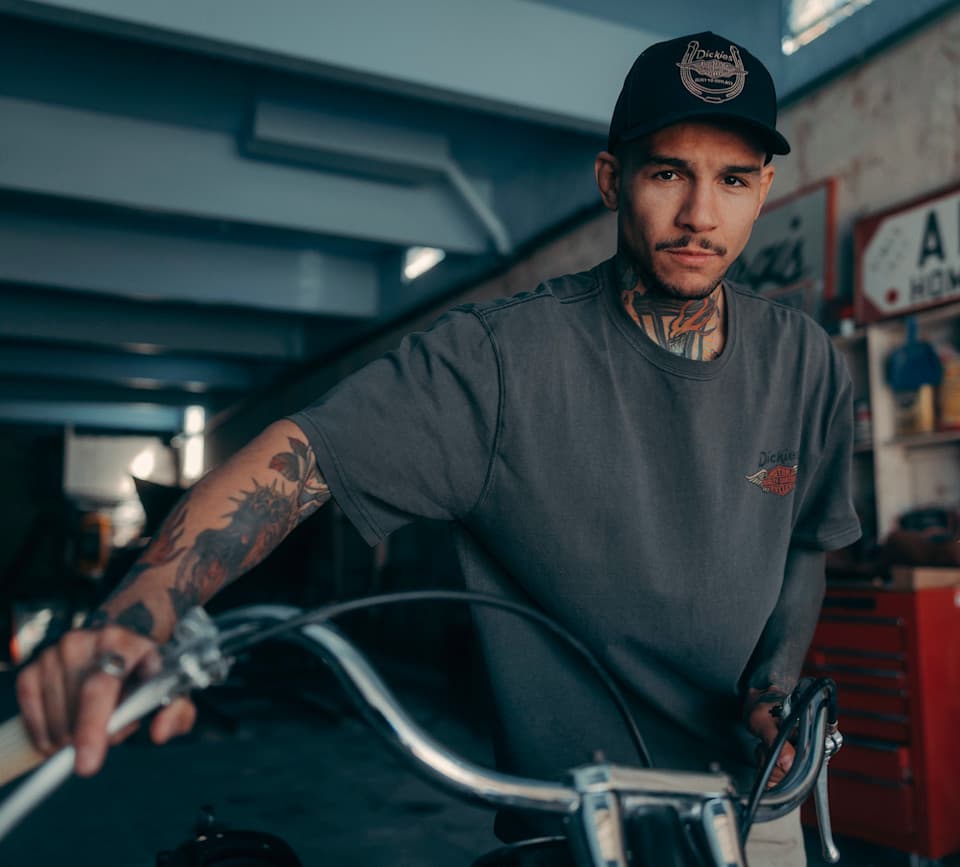 Stile motoc Uomo tatuato con maglietta grigia Dickies e cappellino nero Dickies, in posa su una moto custom. Ambiente officina, stile motociclistico.
