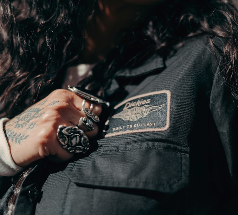 Patch Harley-Davidson e Dickies su giacca, stile autentico Dettaglio di giacca da lavoro Dickies nera con patch Harley-Davidson 'MOTOR CYCLES' alata e scritta 'BUILT TO OUTLAST'. Mano con anelli e tatu