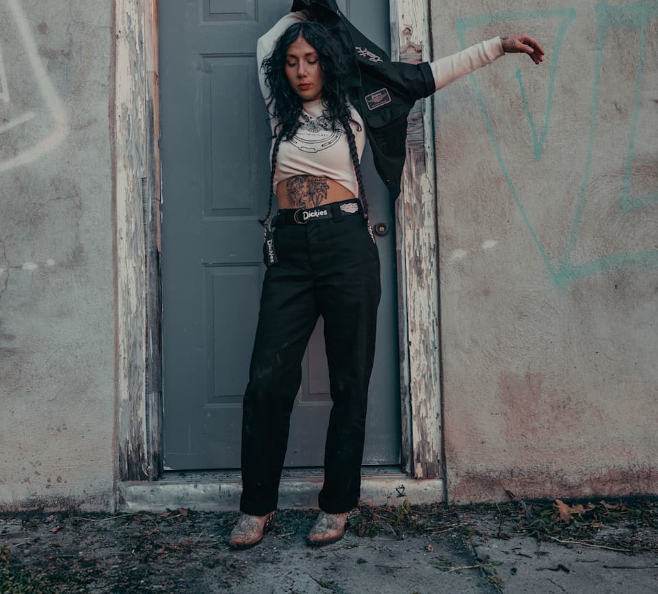 Look workwear e abbigliamento Harley-Davidson Donna con capelli scuri, indossa crop top, pantaloni Dickies neri e giacca da lavoro, con tatuaggi. Posa davanti a porta e muro scrostati, evocando stile abbigliamento moto.