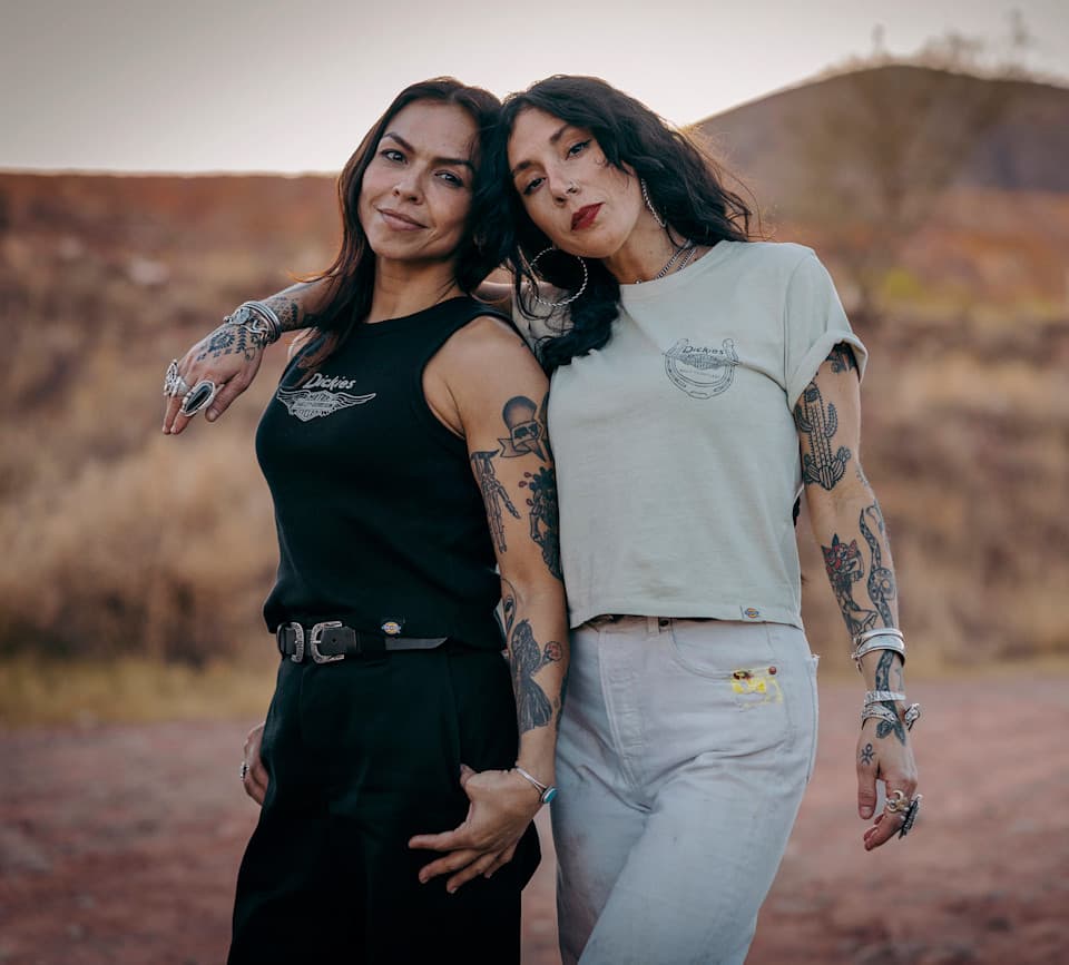Abbigliamento Harley-Davidson: stile autentico e ribelle. Due donne con abbigliamento Dickies, canotta nera e t-shirt chiara, e tatuaggi. Lo stile workwear robusto si allinea al lifestyle Harley-Davidson