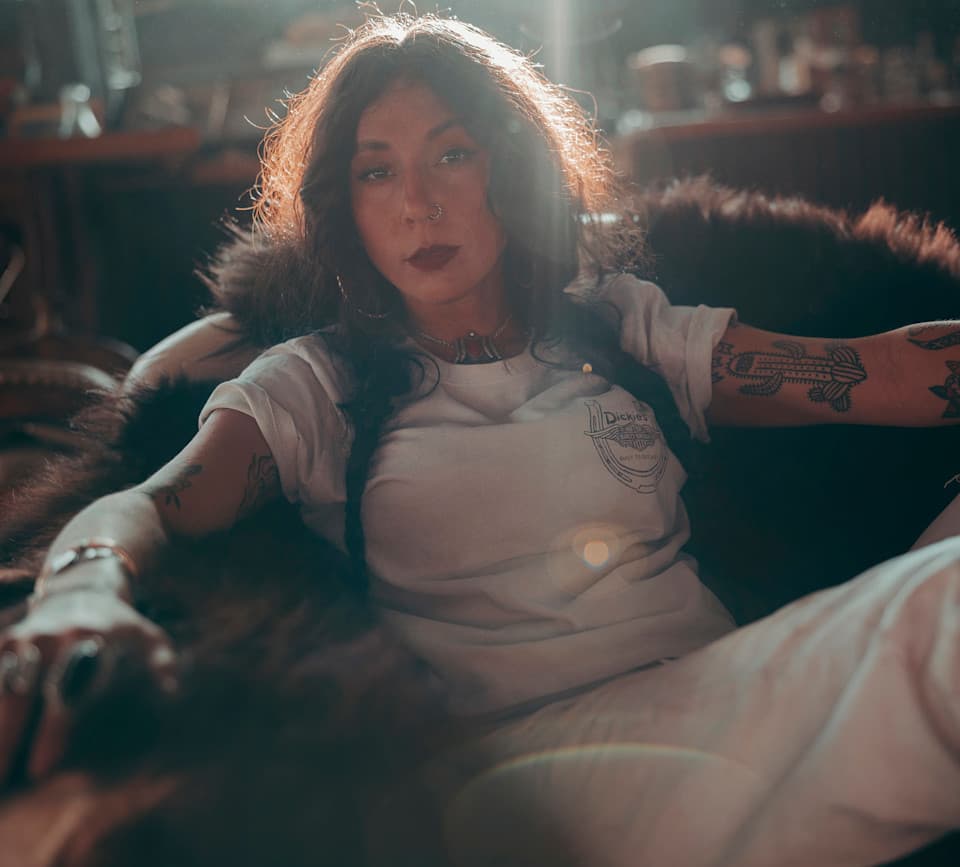 Stile Harley-Davidson: autentico e senza tempo Ritratto di donna con capelli scuri e tatuaggi, indossa una t-shirt bianca Dickies con logo 'Built to Ride' e ferro di cavallo, seduta