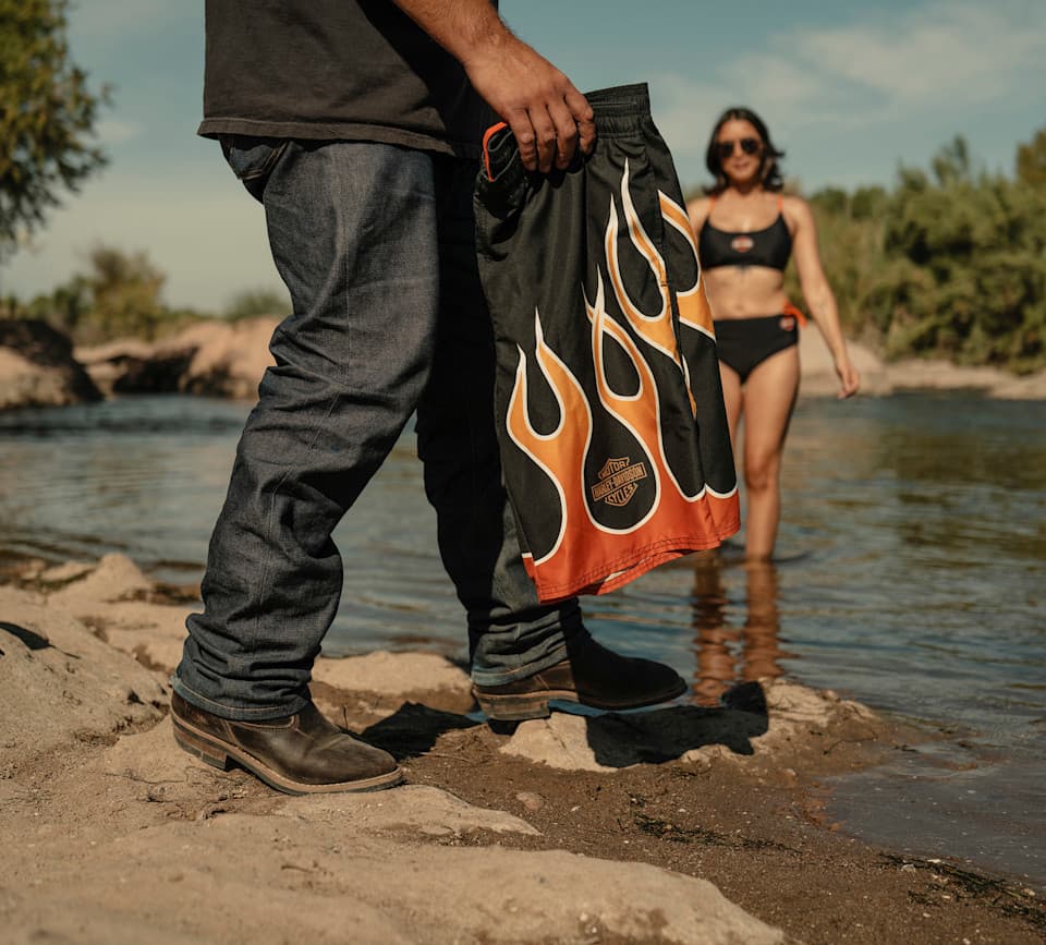Stile Harley-Davidson: abbigliamento mare e tempo libero Uomo in jeans e stivali tiene pantaloncini da bagno neri con fiamme e logo Harley-Davidson. Donna in bikini Harley-Davidson in acqua.