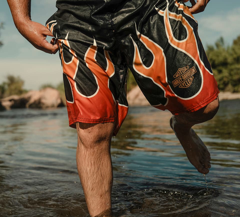 Pantaloncini da bagno Harley-Davidson con fiamme in acqua Uomo in acqua con pantaloncini da bagno neri con fiamme arancioni e rosse e logo Harley-Davidson, mano sinistra sul bordo, piede destro sollevato.
