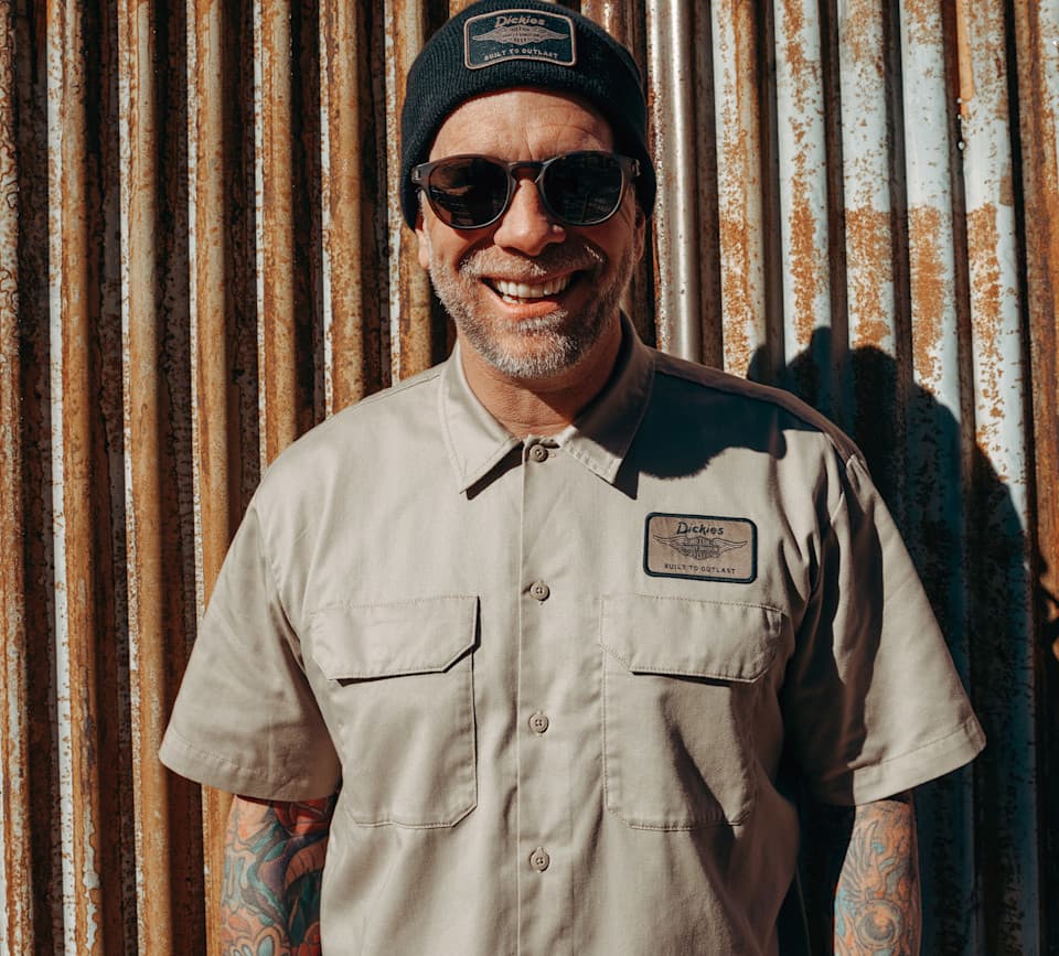 Uomo sorridente con berretto e camicia da lavoro Dickies beige, entrambi con patch 'BUILT TO OUTLAST', occhiali da sole e tatuaggi. Sfondo Uomo sorridente con berretto e camicia da lavoro Dickies beige, entrambi con patch 'BUILT TO OUTLAST', occhiali da sole e tatuaggi. Sfondo